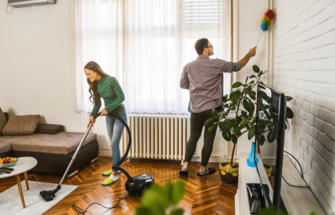  Two people cleaning a bright living room; one vacuuming the floor and the other dusting the wall.