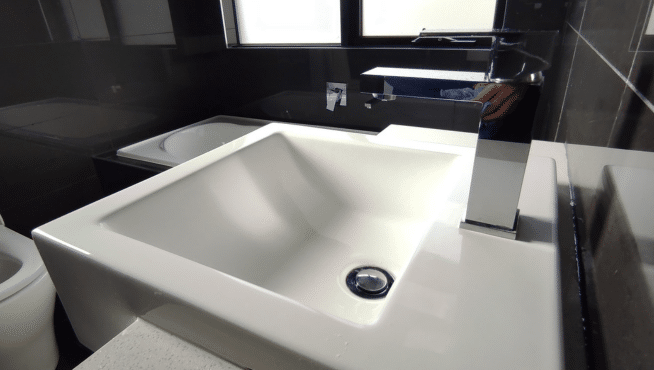 Close-up of a white, square-shaped bathroom sink with a modern stainless steel faucet, set against a dark countertop with reflections visible.