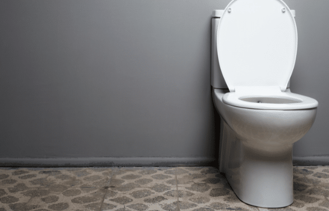 A modern, white toilet against a grey wall with a tiled floor that has a pebble design.