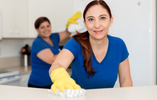 Two smiling professional cleaners working in a kitchen, one wiping the counter and another in the background, symbolizing end-of-lease cleaning services.