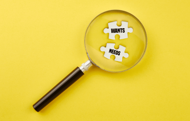 A magnifying glass focusing on jigsaw puzzle pieces labeled "Wants" and "Needs", symbolizing the analysis or assessment of personal or consumer priorities and decision-making.